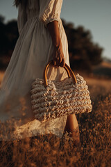 A young woman with long brown hair wears a light dress and holds a crochet bag. She stands in a field during sunset, showcasing a boho summer outfit.