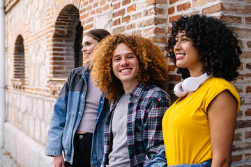 Three cheerful young friends are taking a break from skateboarding, leaning against a brick wall...