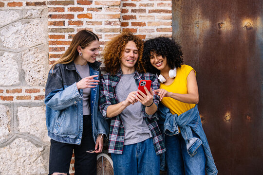 Group of friends looking at a smartphone and smiling, standing next to a brick wall with a skateboard