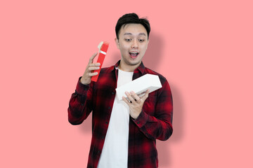 A young Asian man wearing a red plaid shirt over a white t-shirt is holding and looking into an...
