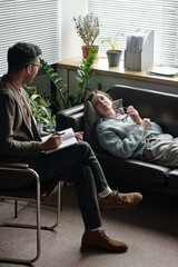 Woman lying on sofa talking to man therapist taking notes during therapy session in office setting...