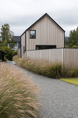 Modern house with wooden exterior surrounded by tall grass and gravel pathway. Overcast sky in the background.