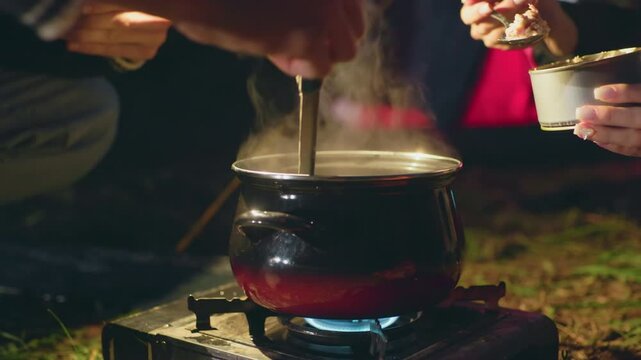 Close up of two people cooking outdoors at night as one stirs boiling pot on gas burner while other adds canned food, with steam rising and warm glow from nearby tent lighting the scene