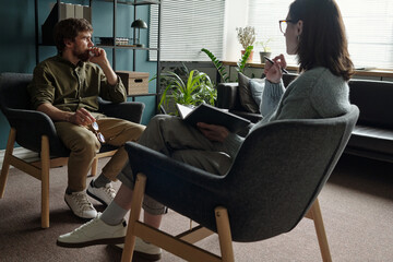 Man sitting in armchair talking to woman therapist holding notebook during therapy session in...
