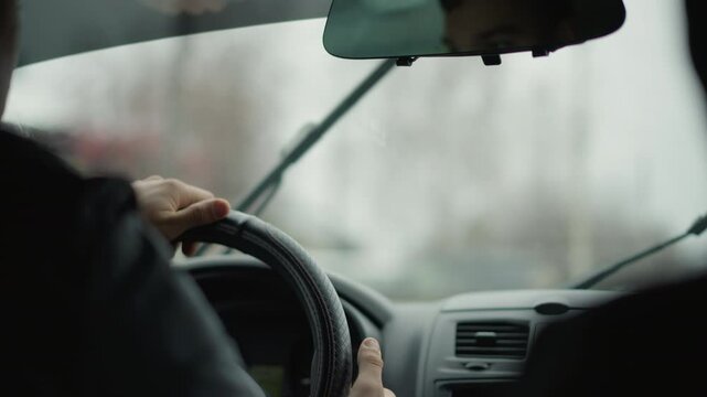 Window cleaner wiping water from front glass while driver navigates rainy urban road, wet windshield and wiper motion visible through raindrops, focus on driver hand on steering wheel and city
