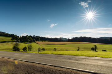 Foothills of the Austrian Alps with asphalt road on foreground and sun on blue morning sky