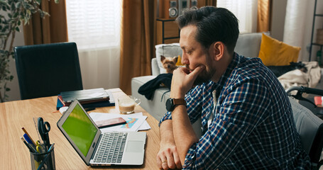 A concentrated man with a disability sits in a wheelchair at his desk, working online. His expression shows determination as he carefully thinks through a digital challenge.