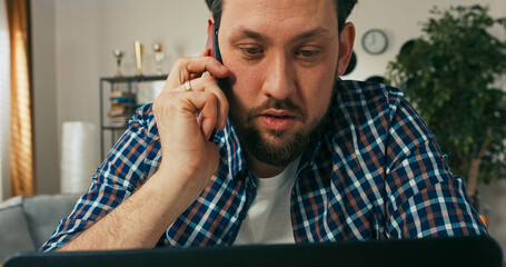 A serious male employee with a beard talks on the phone while working online, confused and thoughtful as he discusses challenges related to a new digital project.