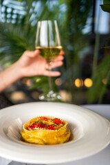 Gourmet ravioli topped with red caviar and herbs on a white plate, with a glass of white wine in hand and a green blurred background. Elegant fine dining close-up.