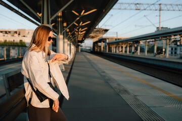 Stylish woman checking time on her watch while waiting for train at sunset, standing on platform of...