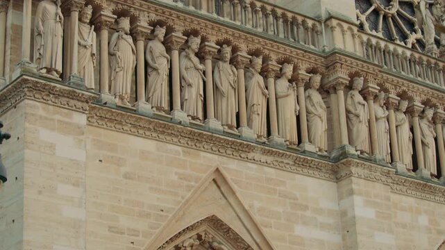 Tilt shot of  the upper fa&ccedil;ade of Notre-Dame Cathedral showing rows of stone saints, ornate gothic arches, rose window, columns and carvings, and partial scaffolding in the background.