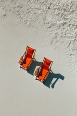 Obraz premium Two orange canvas beach chairs sit side-by-side on a pristine white sand beach, casting shadows in the sunlight