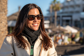 White woman smiling with sunglasses, seated on a terrace during sunset. Warm light highlights her relaxed mood, capturing a joyful summer moment outdoors.

