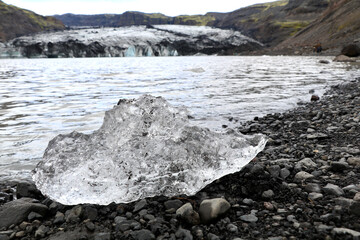 small piece of the Solheimajökull glacier that has broken off and melted into a clear ice diamond and washed ashore on the black sand beach of the glacier lagoon. The glacier in soft focus in the back