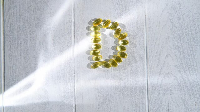 Yellow transparent capsules of vitamin d forming the letter d on a white wooden surface illuminated by sunlight, representing the importance of this vitamin for health and well-being