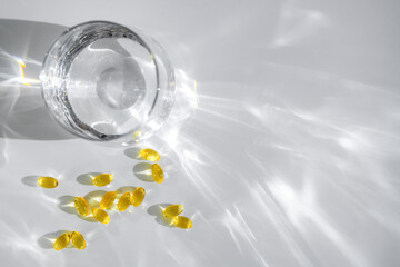 Yellow vitamin D or fish oil capsules are scattered near a glass of water, casting intriguing shadows on a white surface, creating an aesthetically pleasing composition