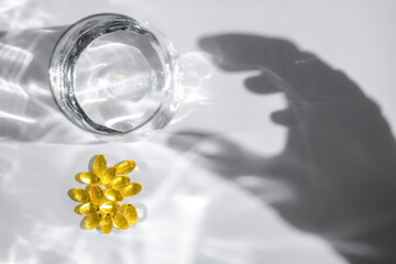 Yellow transparent capsules or pills or vitamins or supplements are near glass of water with hand shadow reaching for them on white background, representing medicine and healthcare