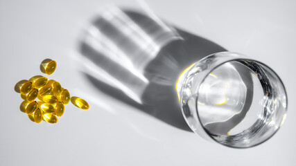 Yellow vitamin D or fish oil capsules are scattered near a glass of water, casting intriguing...