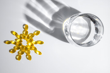 vitamin D or Fish oil yellow transparent capsules forming a sun shape alongside a glass of water create playful shadows on a white background, emphasizing themes of health, wellness, and nutrition