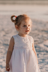 Baby girl in a light dress and two ponytails at sunset on the sea in summer