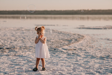 Baby girl in a light dress and two ponytails at sunset on the sea in summer