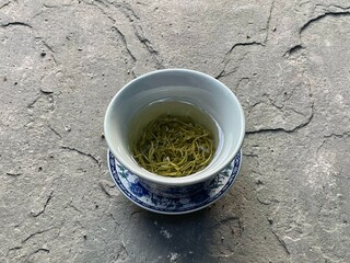 A green tea - filled ceramic cup rests on a blue and white porcelain saucer atop a textured stone...