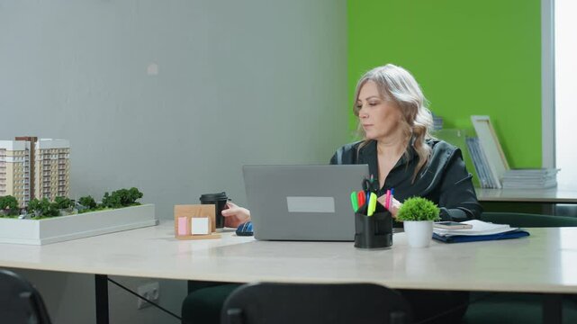 Professional saleswoman pauses work at office desk to sip coffee before resuming typing on laptop, surrounded by building model, folders, and indoor plants in modern workspace