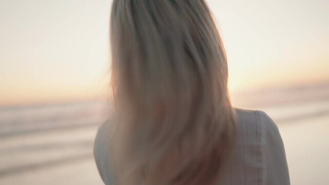 A naturally beautiful young woman with blonde hair and a white dress smiles at the camera during sunset on the beach. She is happy and enjoying life.