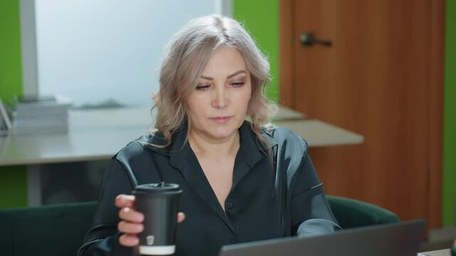 Office assistant pauses while working on laptop to sip coffee before continuing typing, seated in modern office with folders, documents, and natural light creating calm focused work environment