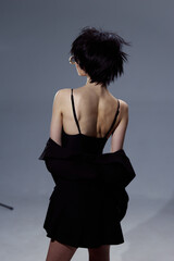 woman with short black hair wearing a black dress and glasses standing with her back against gray...