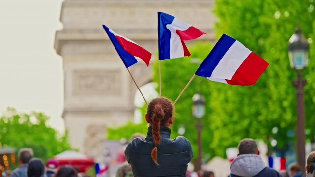 A girl holds three French flags in her hand and waves them on the street against the backdrop of the Arc de Triomphe in Paris during the celebration of the national holiday