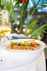 Gourmet salmon tartare with herbs and microgreens, served with toasted bread and white wine on a white plate, set against a lush green outdoor background.