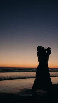 Silhouette of a woman dancing gracefully on the beach as the sun sets. Her movements are fluid and elegant, creating a captivating visual against the backdrop of the ocean and the colorful sky.