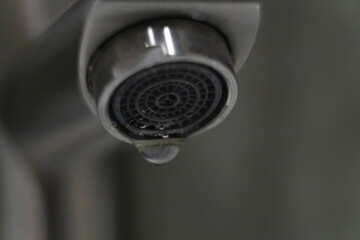 Close-up view of a water droplet poised to fall from a chrome faucet aerator, emphasizing the...