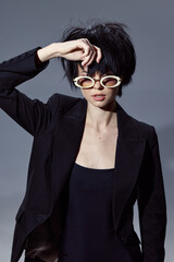 stylish woman wearing black blazer and white round sunglasses posing against gray background with...