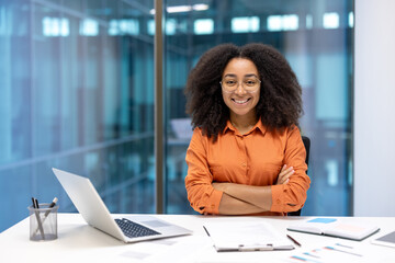 A smiling businesswoman with crossed arms sits in a modern office, radiating confidence and...