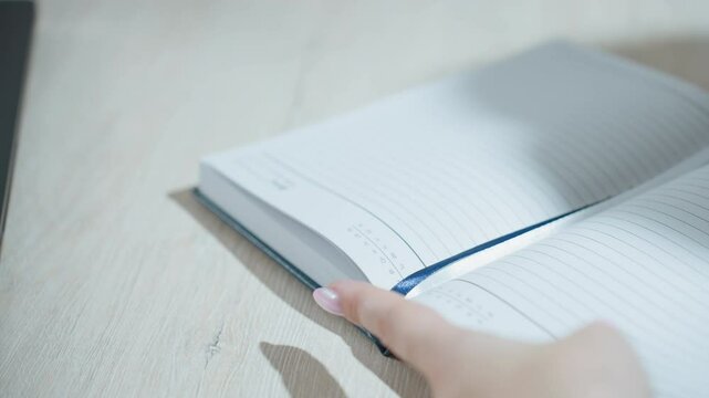 close up of lady with manicured nails flipping open lined notebook on wooden desk, preparing to write with blurred hand motion and ribbon bookmark
