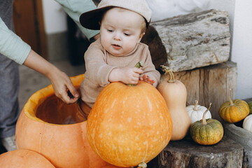 Sweet happy baby boy playing in big pumpkin at front door with autumn rustic decor. Happy halloween and Thanksgiving. Adorable little child in sitting with pumpkins on porch