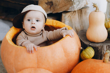 Cute baby boy sitting in big pumpkin at front door. Happy halloween and Thanksgiving. Adorable little child in cozy clothes sitting with pumpkins on porch, autumn decor at home