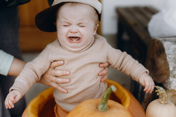 Cute baby boy crying in big pumpkin at front door. Sad emotional little child sitting with pumpkins on porch, autumn decor