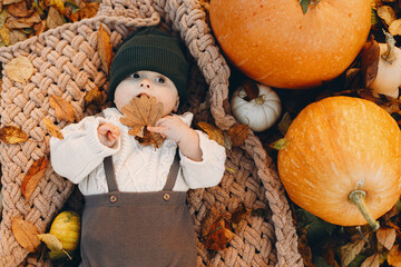Adorable baby boy lying on cozy blanket with pumpkin and autumn leaves, top view. Cute happy little...