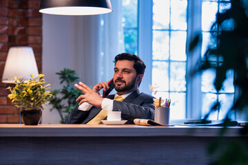 Indian businessman talks on phone while enjoying tea, showing relaxed yet connected office routine