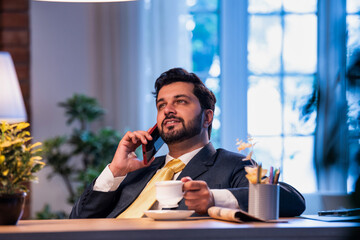 Indian businessman talks on phone while enjoying tea, showing relaxed yet connected office routine
