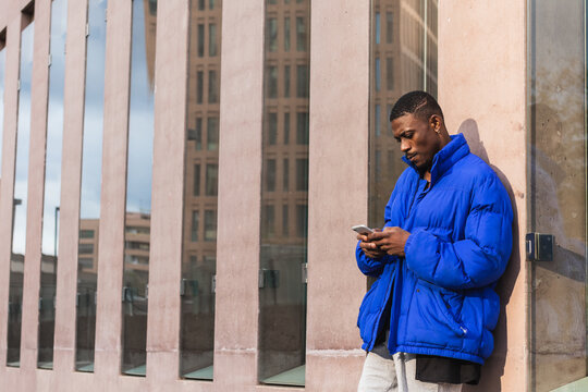 Cool young African Black man hipster or tourist wearing blue jacket standing on city street outdoors holding smartphone using mobile cell phone technology smartphone device - Powered by Adobe