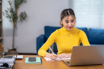A woman in a yellow sweater is sitting at a desk with a laptop and a notebook