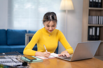 A woman in a yellow shirt is sitting at a desk with a laptop and a notebook
