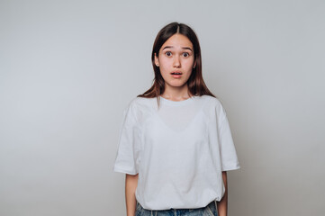 Surprised young woman with neutral expression standing against light background in casual attire