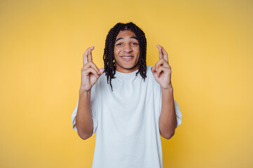 Young african man with braided hair shows crossed fingers for luck in front of yellow background...