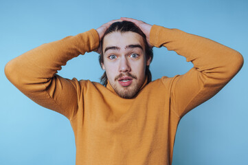 Young man expressing shock in a bright sweater against a blue background while holding his head in...