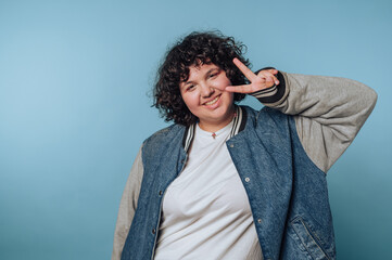 Person showing peace sign in a casual outfit against a light blue background in an indoor setting